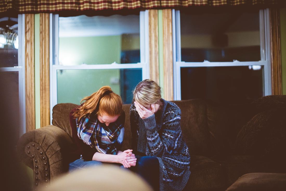 Two women sitting on a couch indoors, looking down as if upset.