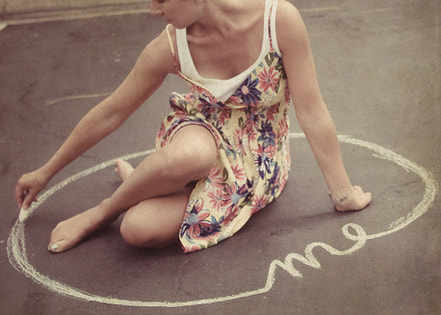 A photograph of a woman sitting on the ground outside, drawing a circle around her and the word 