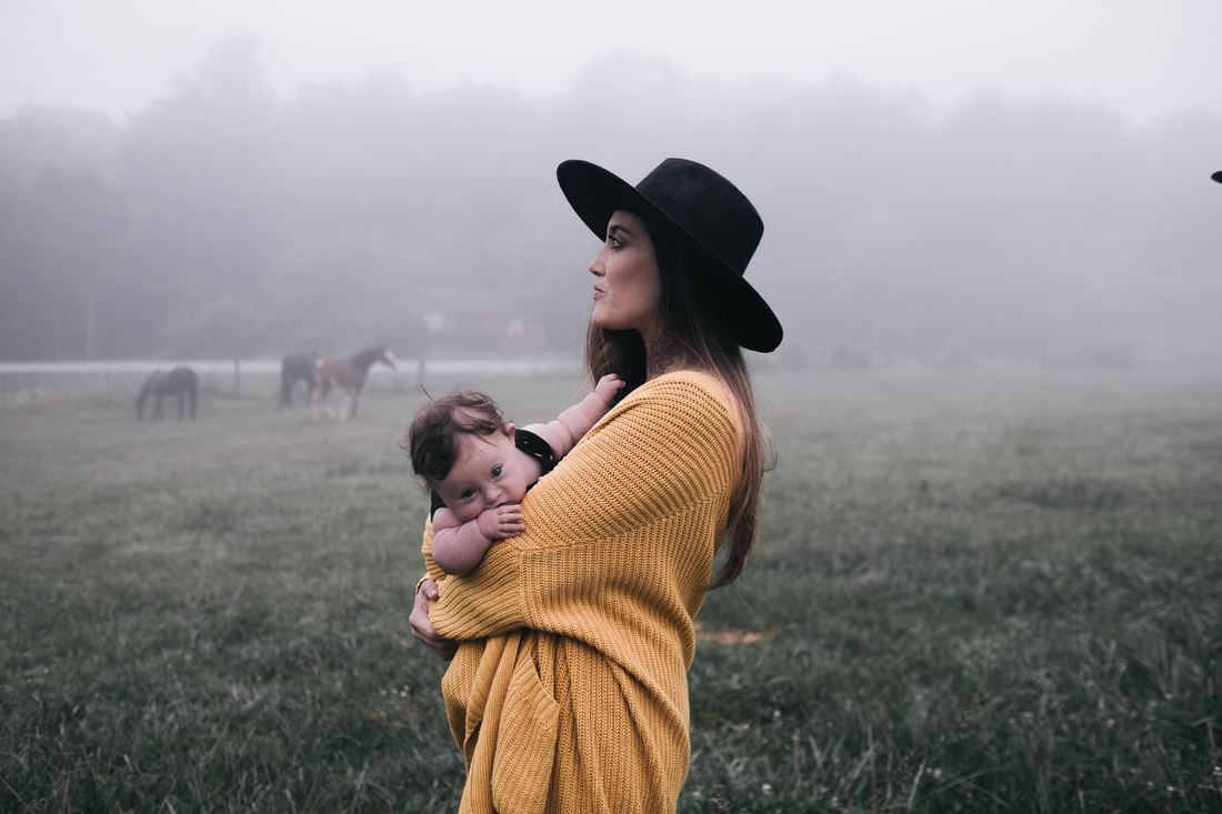 A photo of a woman holding a baby while she stands outside, with horses in the background.
